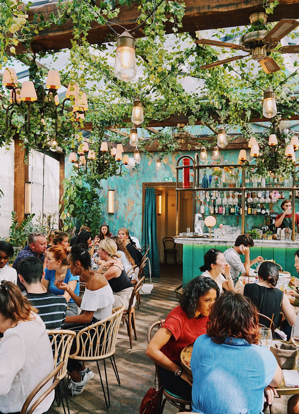 Busy restaurant with large bar, ceiling beams covered with greenery, large string lights, and busy tables.